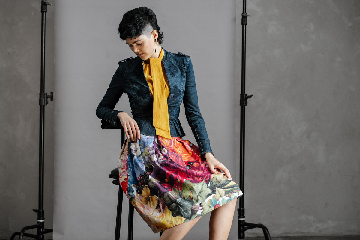 Woman sitting on a stool wearing a fitted denim jacket and yellow top, with the by Audrey & Grace, Zero Waste Big Bloom Skirt, and red shoes against a gray background.