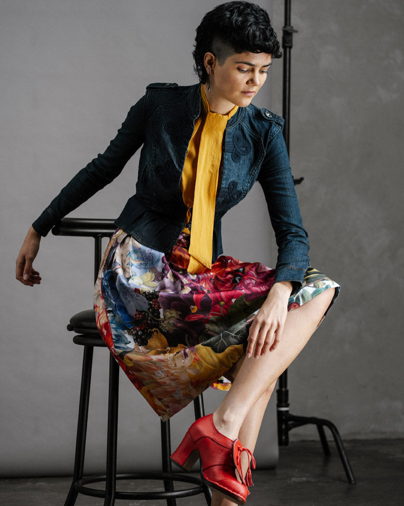 Woman sitting on a stool wearing a fitted denim jacket with the by Audrey & Grace, Zero Waste Big Bloom Skirt, and red shoes against a gray background.