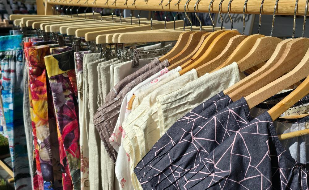 A wooden clothes rack with Zero Waste tops in the foreground. The Organic cotton fabric shows charcoal background with soft pink abstract lines and Big Bloom and Ocean Zero Waste Skirts in the background. Australian made zero waste clothing