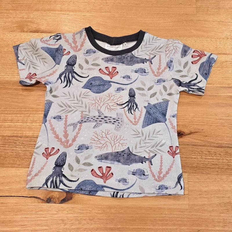 We can see the front of an Australian made childs tee shirt, as a flat lay featuring an underwater scene print with stingray, squid, hammerhead shark and more.  Featuring inky blue, red, peach and grey colours
