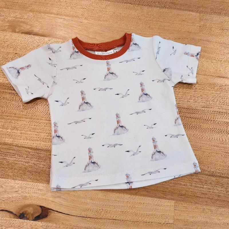 A childrens t-shirt with a lighthouse and seagull print on a crisp white background.  Finished with a rust coloured ribbing around the neck