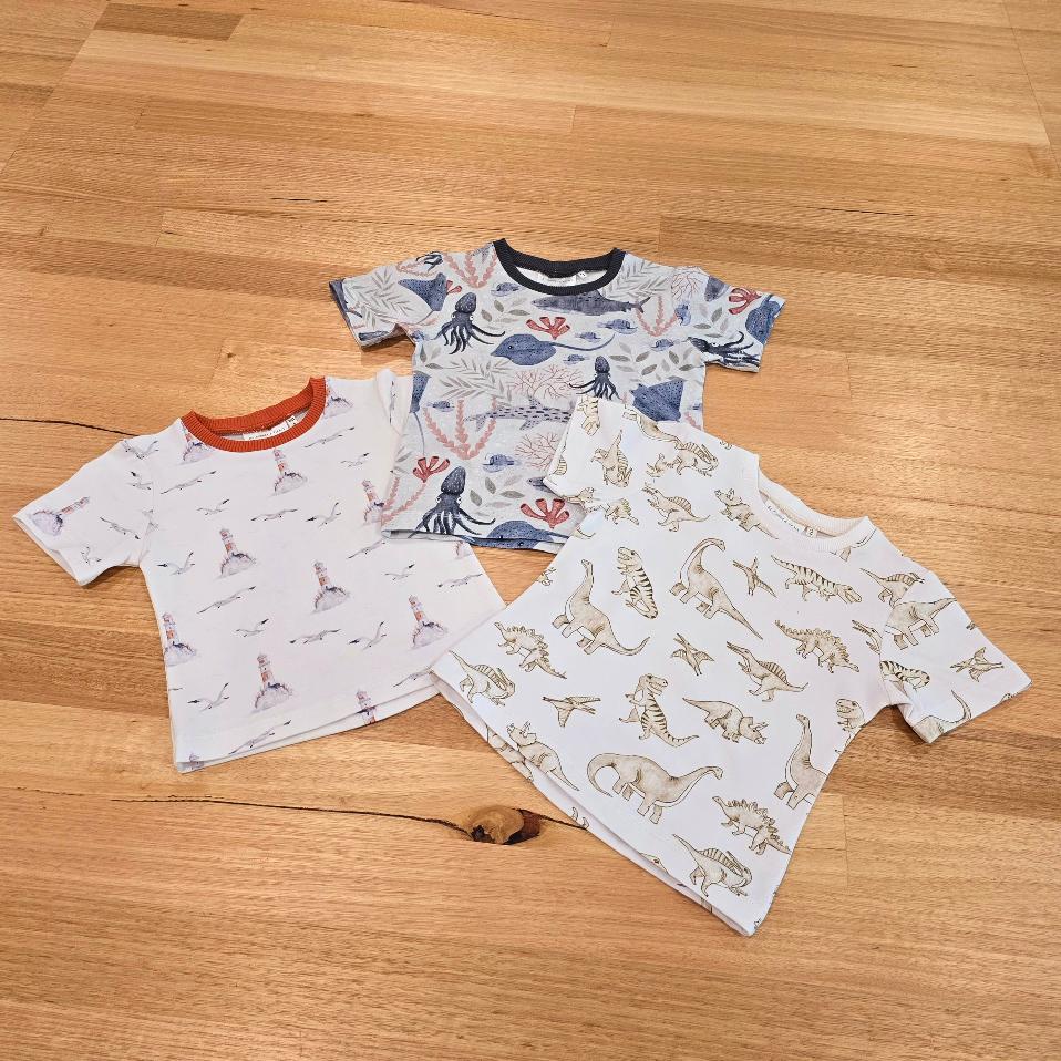 Flatlay showing three Australian made childrens t shirts. On the botton to the left is a tee with lighthouses and seagulls, in the middle is an underwater scene with stingrays, hammerhead sharks and squid, and the third tee, on the right features monochrome dinosaurs.