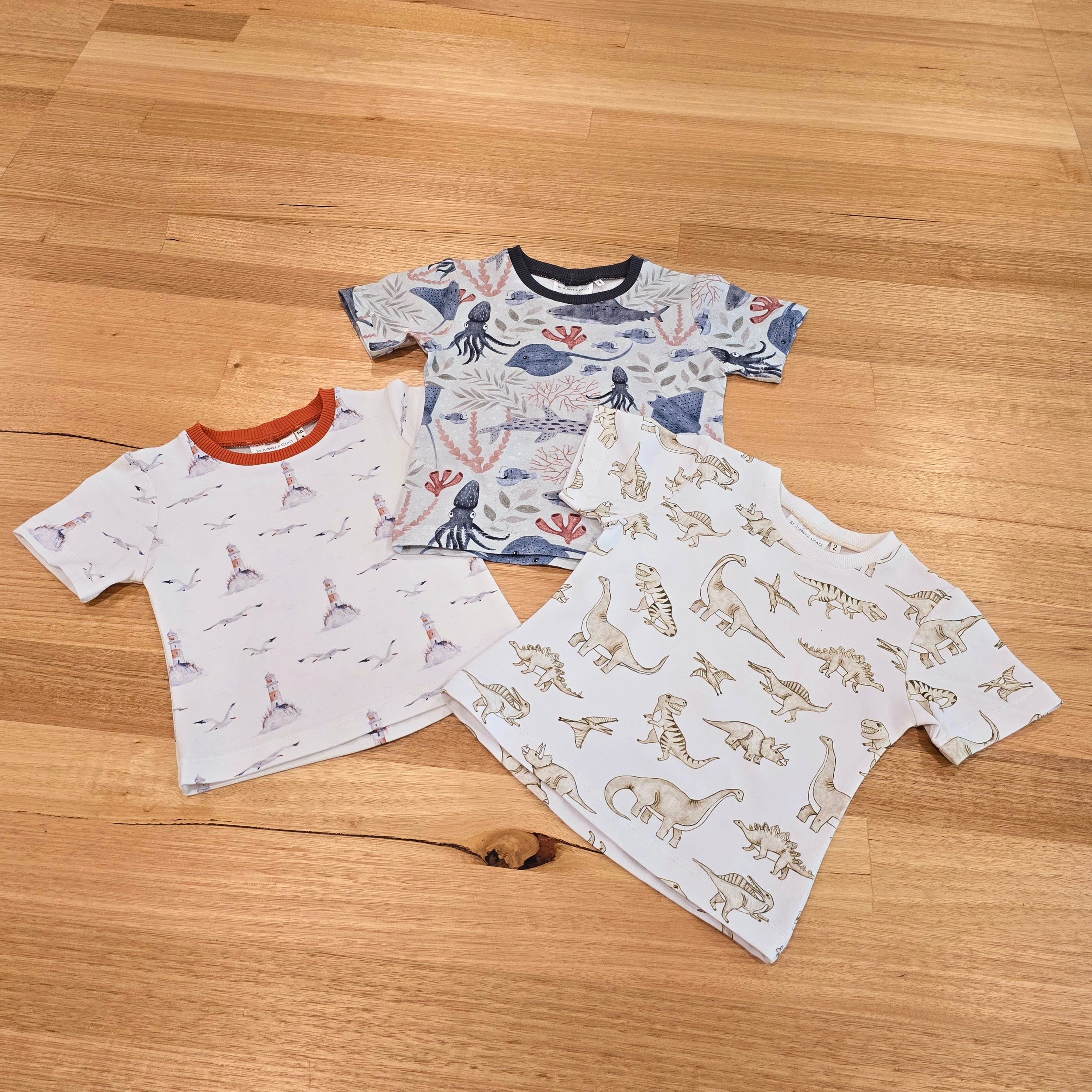 Flatlay showing three Australian made childrens t shirts. On the botton to the left is a tee with lighthouses and seagulls, in the middle is an underwater scene with stingrays, hammerhead sharks and squid, and the third tee, on the right features monochrome dinosaurs.