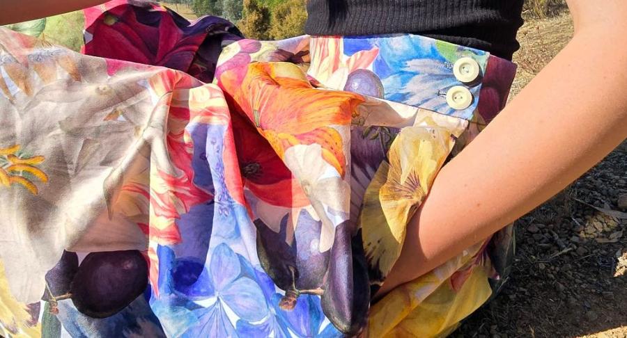 Model sitting on the ground wearing the Zero Waste Big Bloom skirt with her hand in the almost invisible, large side seam pocket.  2 natural corozo buttons in off white can be seen on the deep waistband. Large format flowers in yellow, blue, indigo, deep red and white can be seen printed on the organic cotton fabric of this stunning Australian made womens skirt.