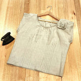 Flat lay of a by Audrey and Grace Zero Waste top in sand colour, on a wooden table with a small, vintage iron next to it.  There are vertical pin tucks across the front of the top and short ones at the sleeve edges and at neckline. A rosette can be seen at the front  of the left shoulder.