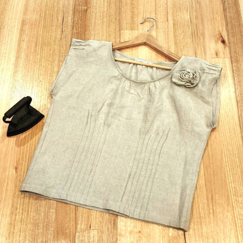 Flat lay of a by Audrey and Grace Zero Waste top in sand colour, on a wooden table with a small, vintage iron next to it.  There are vertical pin tucks across the front of the top and short ones at the sleeve edges and at neckline. A rosette can be seen at the front  of the left shoulder.