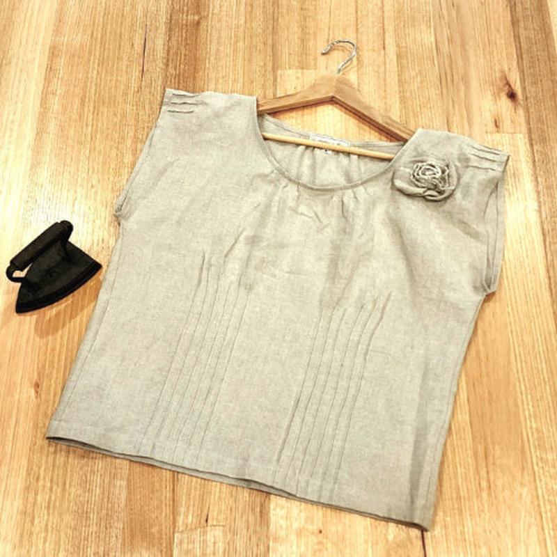 Flat lay of a by Audrey and Grace Zero Waste top in sand colour, on a wooden table with a small, vintage iron next to it.  There are vertical pin tucks across the front of the top and short ones at the sleeve edges and at neckline. A rosette can be seen at the front  of the left shoulder.