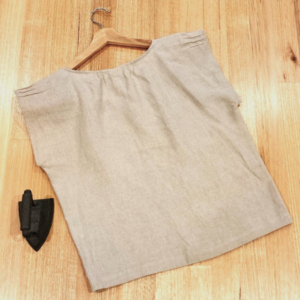 Back view flat lay of a by Audrey and Grace Zero Waste top in sand colour, on a wooden table with a small, vintage iron beside it.  There are short, decorative pin tucks at the sleeve edges and at neckline. 