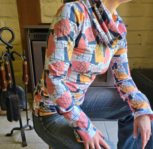 Model wears the Ruby Jumper in the Jumble print. Sitting side on , showing long boddy and long sleeves with foldable cuffs at full length and cowl turtle neck collar softly folding around neck