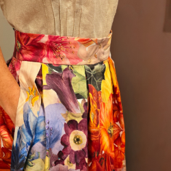 We see a model wearing the Australian made by Audrey and Grace Zero Waste Big Bloom Box Pleat Skirt which is covered in giant, beautifully coloured flowers including orange Hibiscus, passion fruit flower pink and purple Fuscias and more. The model is standing against a dusty purple wall background with her right hand in her side pocket and a wide waistband above. The 100% organic cotton fabric softly hangs from perfect large box pleats