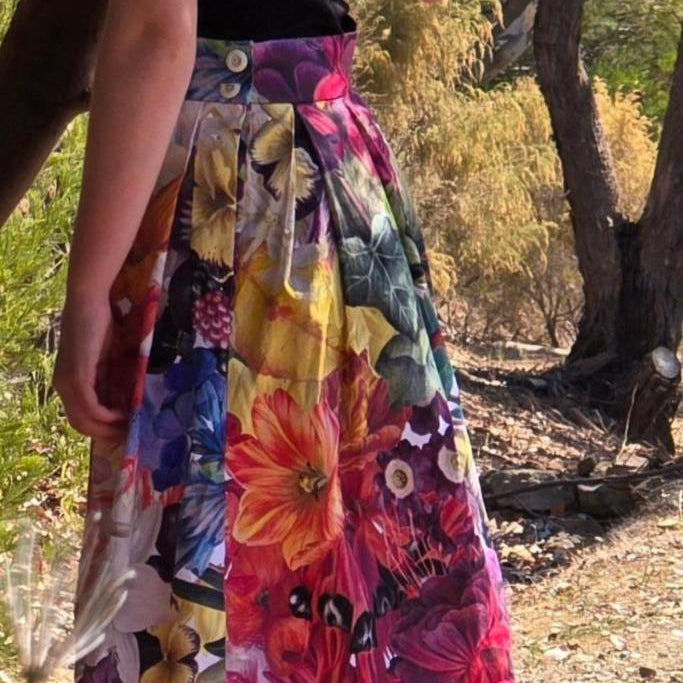 A model is standing with her back to us, with one arm bent up and leaning against a tree branch, with trees and brush in the background. She is wearing the by Audrey and Grace Zero Waste Big Bloom Box Pleat Skirt which is covered in giant, beautifully coloured flowers including a white Lily, orange Hibiscus, a Sturt's Desert Pea, pink and purple Fuscias and more. A wide waistband has two natural corozo buttons fastening it at the side. The 100% organic cotton fabric softly hangs from the box pleats.