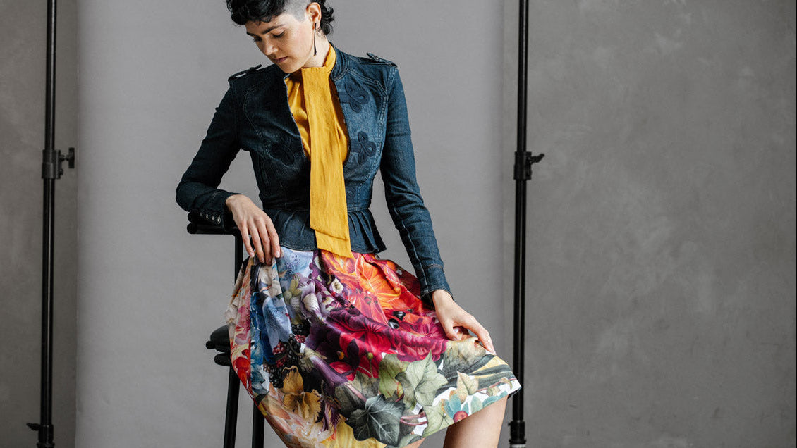 Woman sitting on a stool wearing a fitted denim jacket and yellow top, with the by Audrey & Grace, Zero Waste Big Bloom Skirt, and red shoes against a gray background.