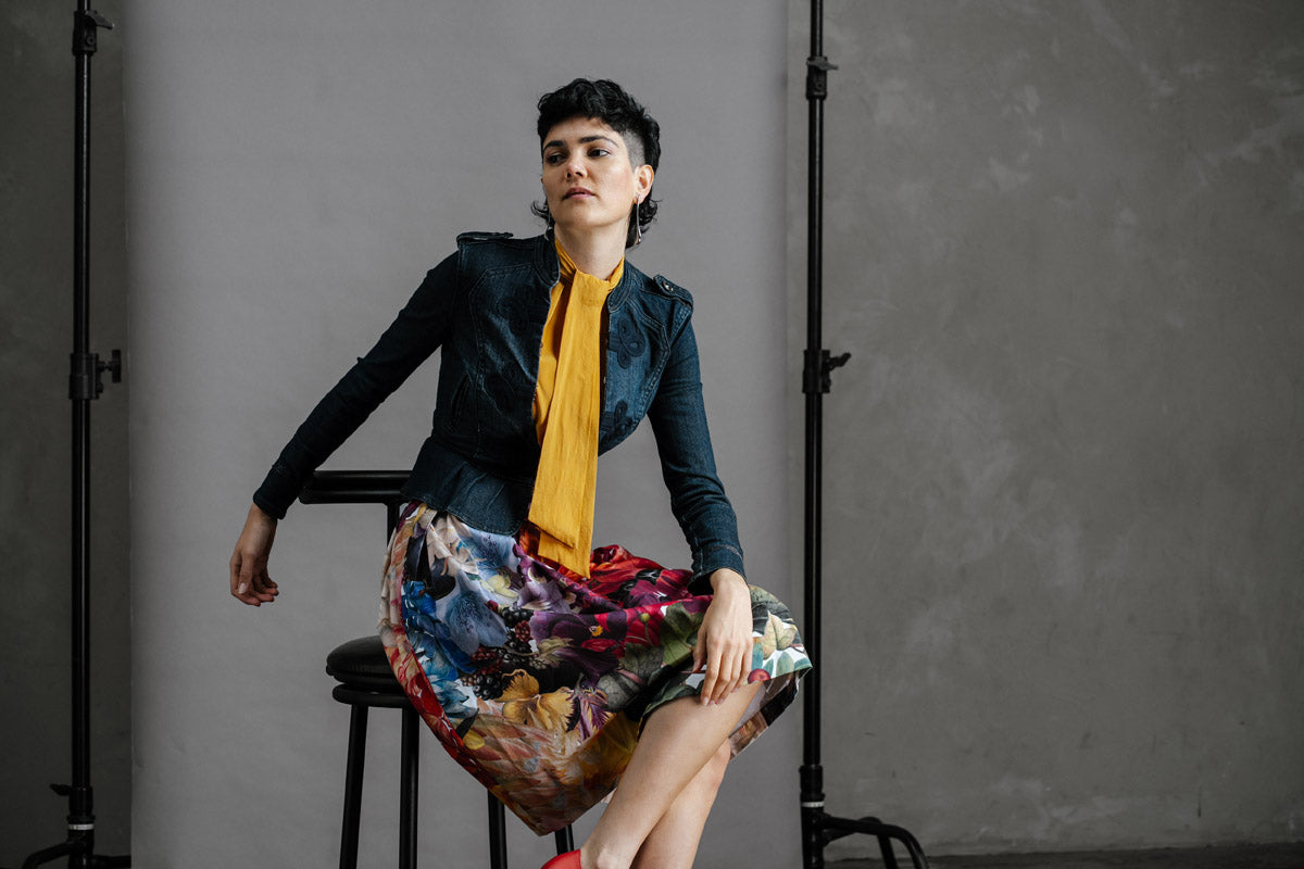 Person sitting on a stool wearing a dark jacket, yellow shirt, and the Big Bloom Zero Waste skirt against a grey background.