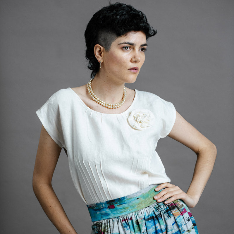 Woman wearing an oyster coloured dress top, a zero waste top from by Audrey and Grace, featuring pin tucks from waist up to bust line, around the neck and on the sleeves, with a round neck line and a flower from self fabric at the front of the left shoulder.