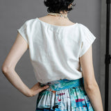 Woman facing the back, wearing pearls around her neck and an oyster coloured dress top with short cap sleeves, a zero waste top from by Audrey and Grace, featuring pin tucks from waist up to bust line, around the neck and on the sleeves, with a round neck line and a flower from self fabric at the front of the left shoulder.