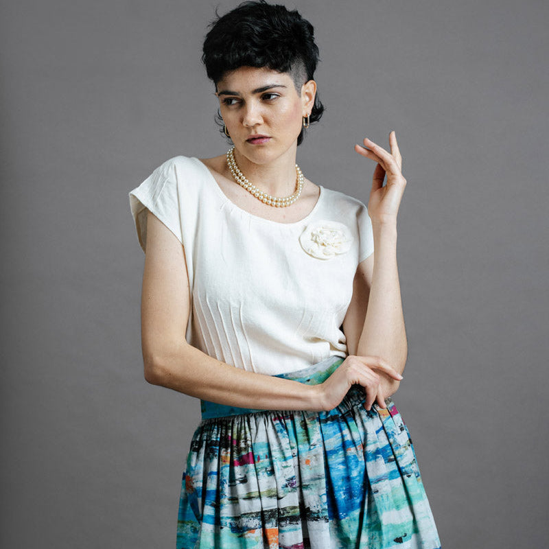 3/4 length image of woman wearing the Zero waste oyster top with the Zero Waste Ocean skirt. She has one arm folded across her body, touching the tip of her other shoulder, with the other hand raised towards her face.  She has short cut dark hair and is wearing a pearl chocker.