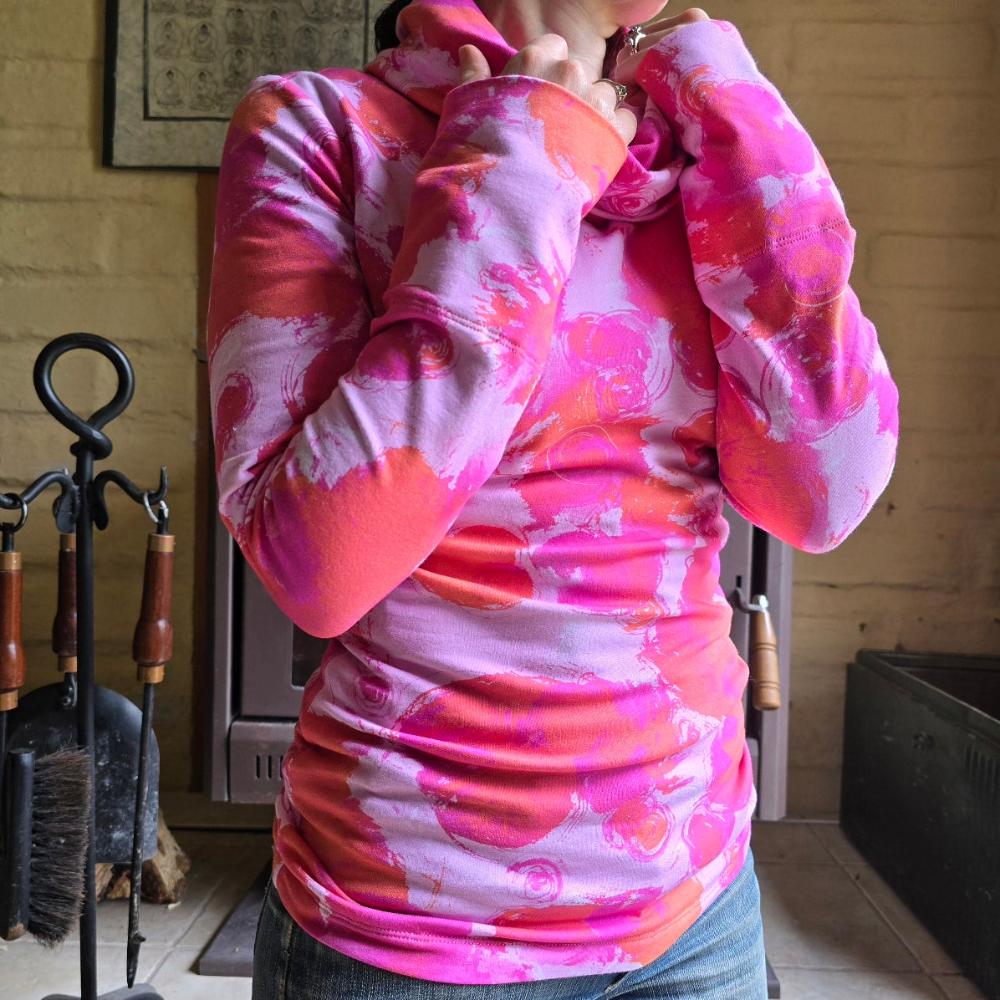 Model wearing the Ruby Jumper Fairy Floss with fuscia pink and orange painted circles on light pink base. This Australian Made womans jumper is a slim fit on the model, who's hands are holding the large, turned over collar and is standing on an angle