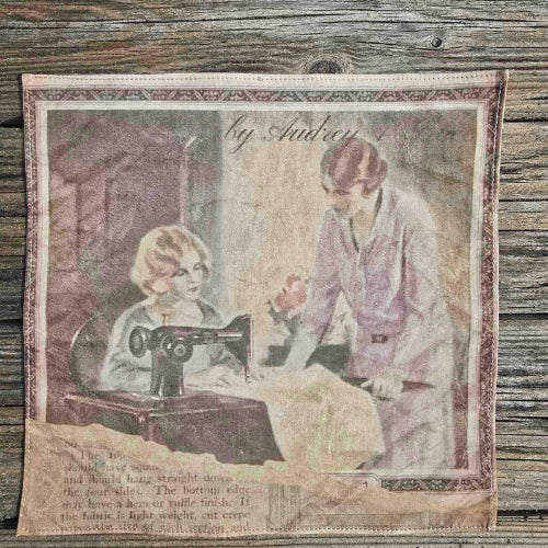 The image shows a woman's handkerchief depicting a scene with two women at a 1920s Singer sewing machine. One is sewing, and the other is leaning over and instructing, wearing a smart Mauve skirt suit. The image appears torn, with what appears to be a page of text, well aged, underneath.