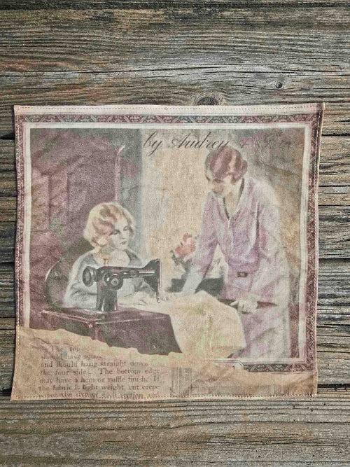 The image shows a woman's handkerchief depicting a scene with two women at a 1920s Singer sewing machine. One is sewing, and the other is leaning over and instructing, wearing a smart Mauve skirt suit. The image appears torn, with what appears to be a page of text, well aged, underneath.