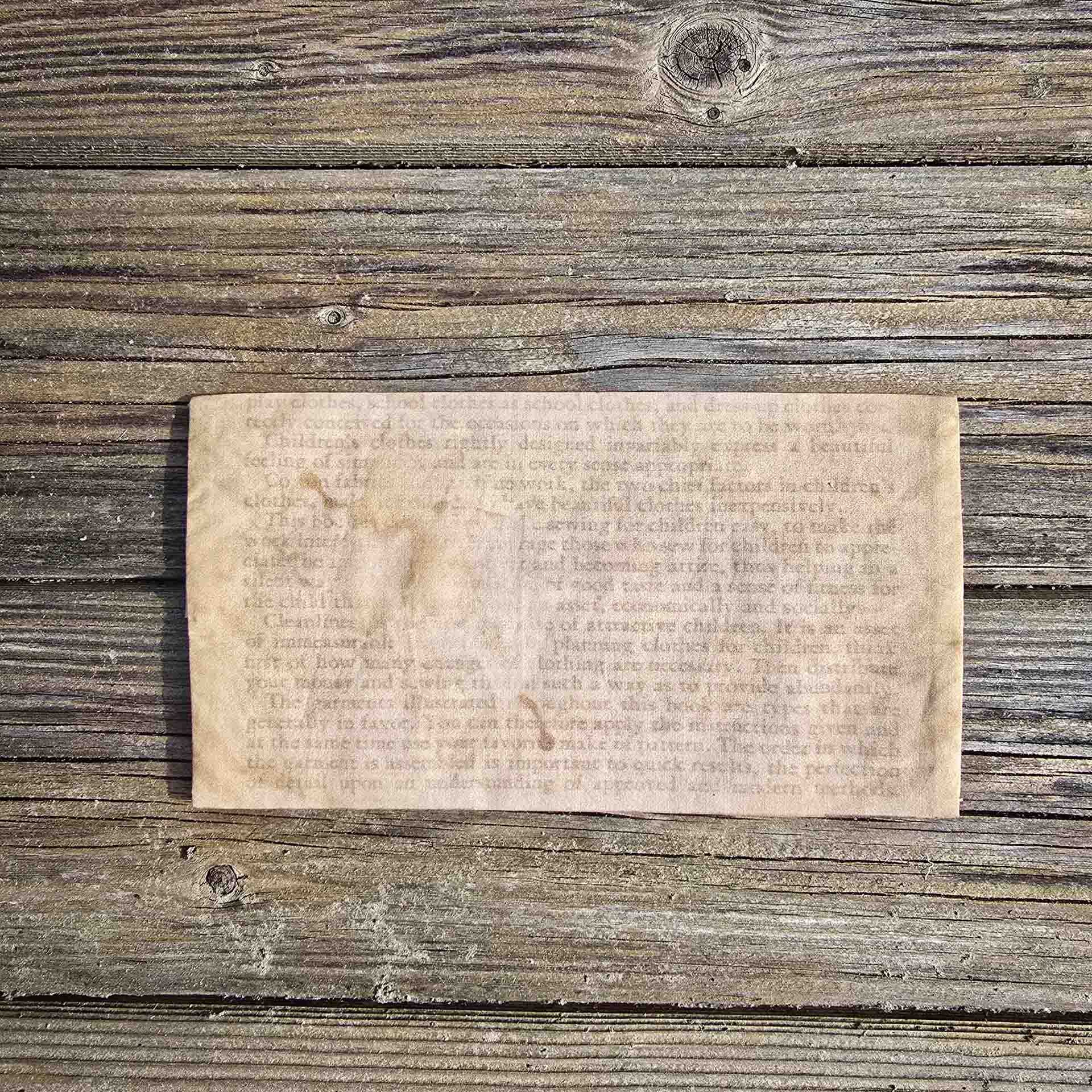 The back side of a delicate rectangular hanky pouch, showing text from a booklet, hard to read, aged with a coffee stain over the top of it.