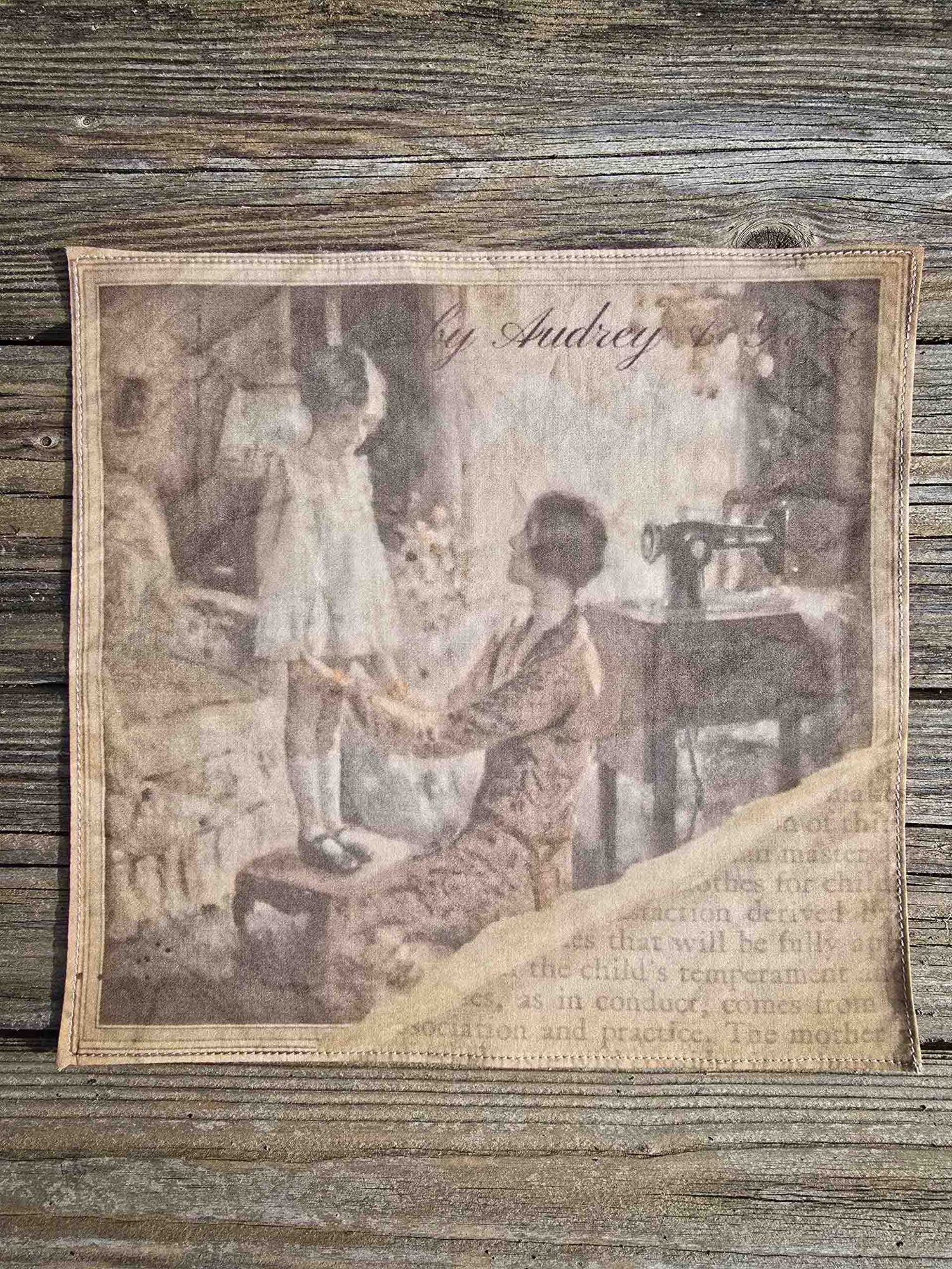 A ladies handkerchief with a special print, depicting a 1920s mother checking the hem length of her littles girls dress as she stands on a stool, with her sewing machine in the background. The imae shows a torn edge with a page of text visible eneath, with an aged feel to the image.