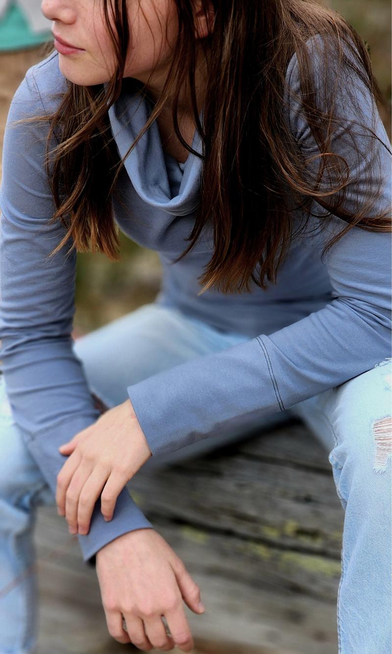 The Ruby Jumper in Storm, a blue grey colour, with large cowl neck, Australian Women's Jumpers made from Australian cotton — model sitting on a log and leaning forward