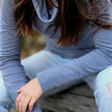 The Ruby Jumper in Storm, a blue grey colour, with large cowl neck, Australian Women's Jumpers made from Australian cotton — model sitting on a log and leaning forward