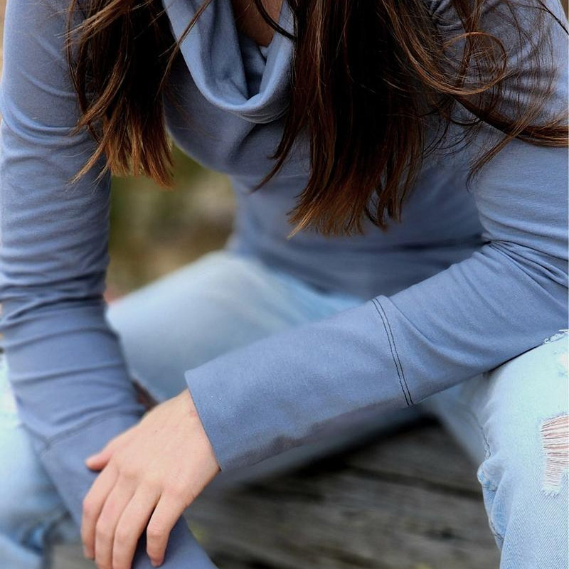 The Ruby Jumper in Storm, a blue grey colour, with large cowl neck, Australian Women's Jumpers made from Australian cotton — model sitting on a log and leaning forward