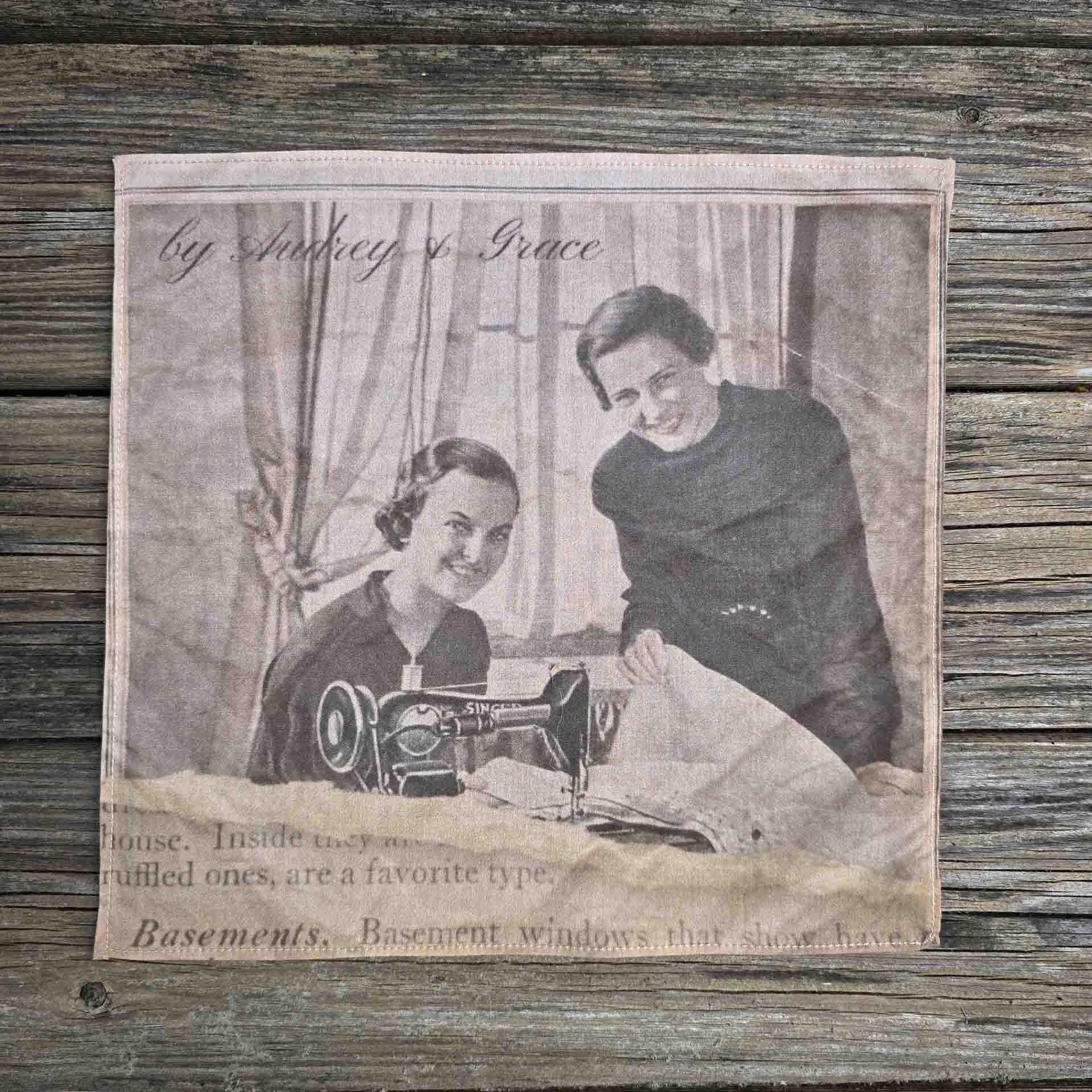 Image of a ladies handkerchief with the image f two women at a Singer sewing machine smiling at the camera. They are framed by delicate drapes in the background and one is holding up some fabric. The image appears to be torn with a page of aged text underneath.