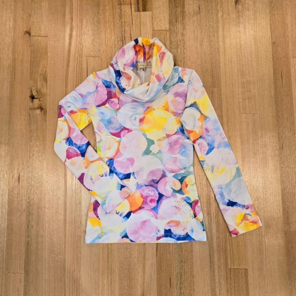 The Ruby Jumper with large cowl turtle neck collar and long sleeves with reversible cuff, in bubbles print which has a white base with overlapping, non uniform watercolour circles,  in colours of dark blue, pinks, soft yellow, mauve and teal green, all with different soft opacities. One arm is straight and just away from he right side of the body and the left sleeve is folded back towards the body
