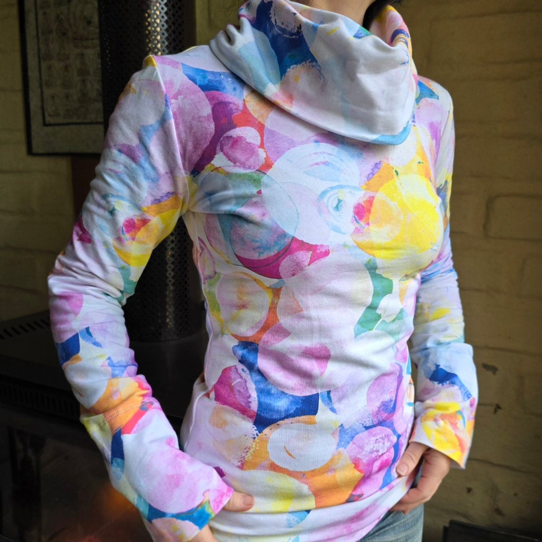The Ruby Jumper with Large cosy Cowl Turtleneck folded over loosely. Jumper being worn by a model who is standing in front of a mud brick wall and just the torso can be seen, angled towards the right of the image.  The model is holding the bottom of the jumper and has the long sleeves covering the tops of her hands. The print has a white base with overlapping, non uniform circles in colours of dark blue, pinks, soft yellow, mauve and teal green, all with different soft opacities and a watercolour style.