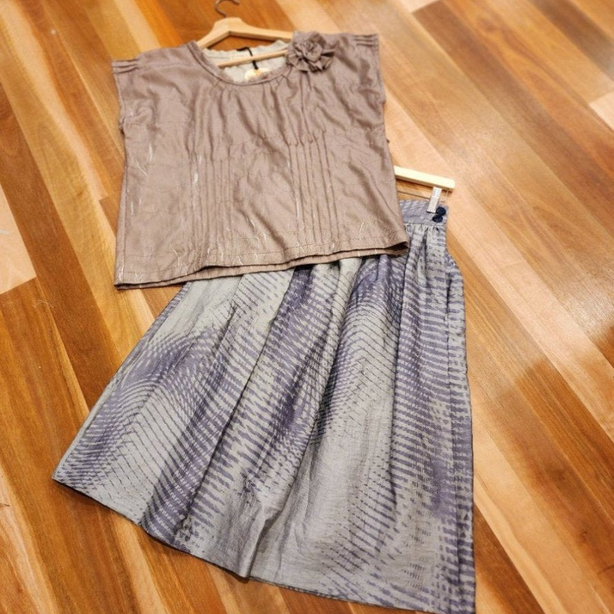 A flat lay showing the by Audrey and Grace zero waste skirt with an abstract wave print lane flat on wooden boards on a wooden coat hanger with a by Audrey & Grace zero waste top also on a  coat hanger laying over the top of the skirts top left corner. Blue vintage buttons can be seen on the deep waistband of the Australian Made skirt. The Australian made top features a rosette at the left shoulder and is made from the offcuts of the shirt and there are featured pin tucks at the front, sleeves and neckline